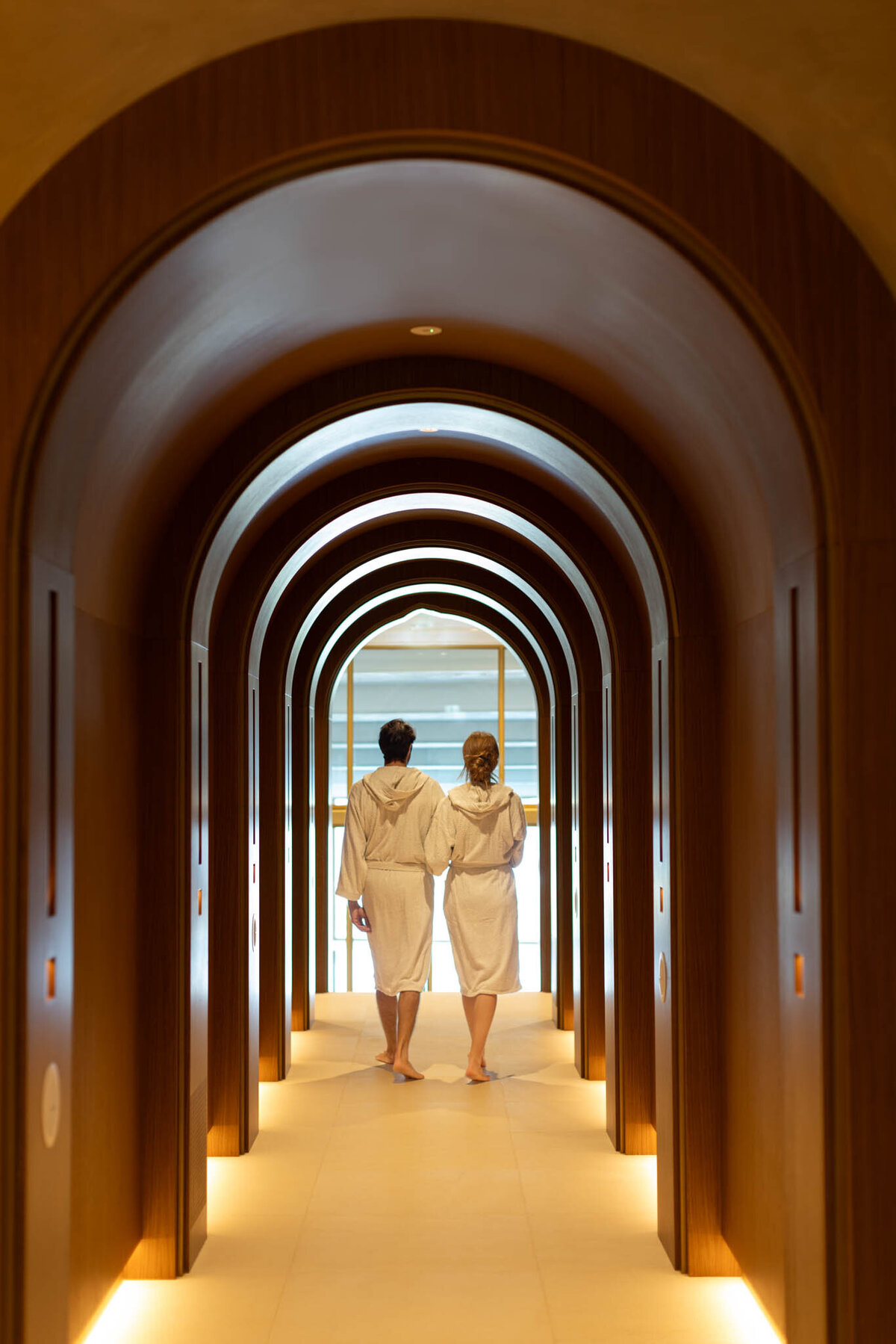 A couple in white robes walk hand in hand down a warmly lit hallway with arched wooden doorways. Their backs are to the camera, leading towards a bright, inviting area at the end of the corridor.