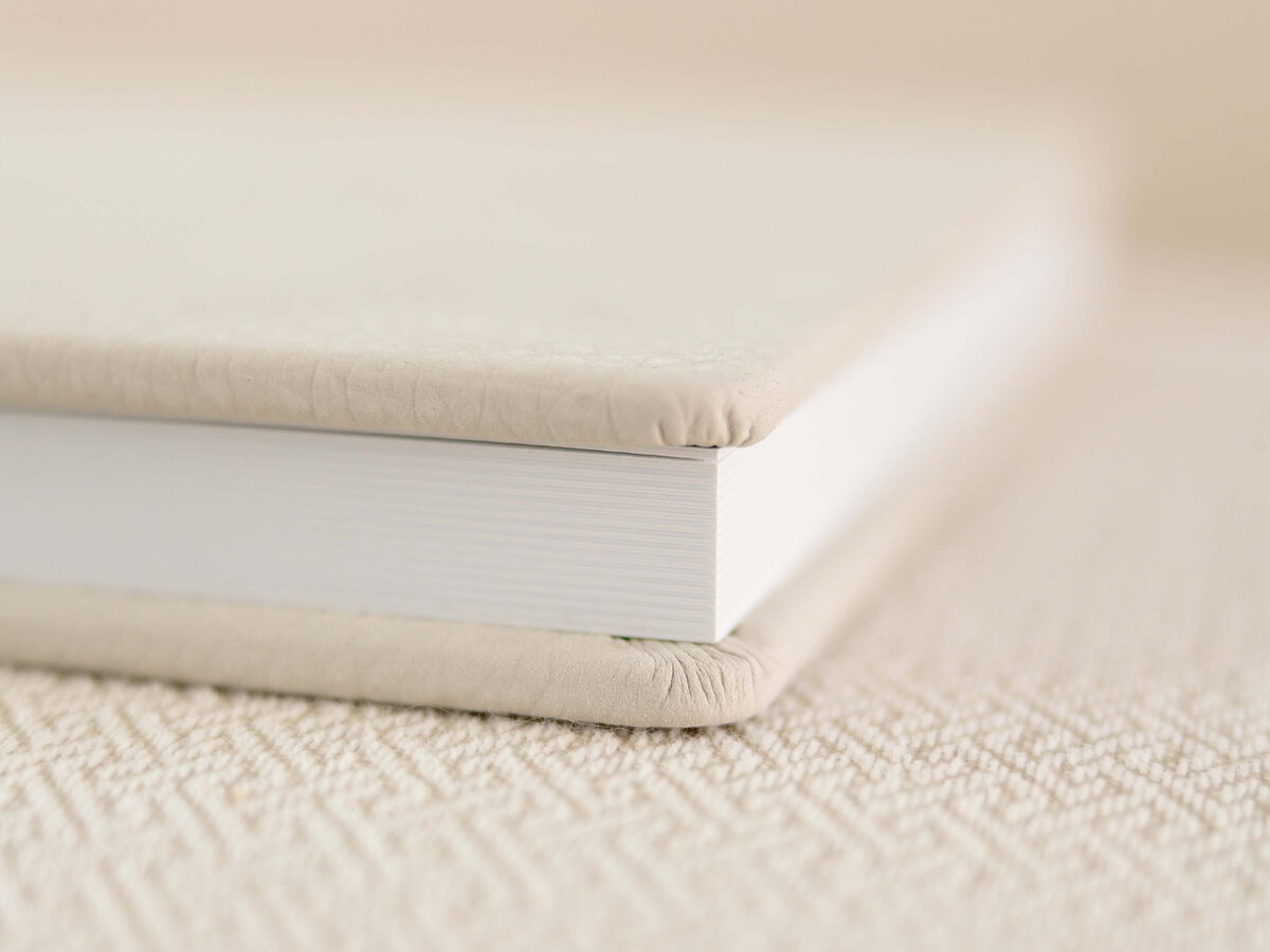 Close-up of a closed, hardcover book with a beige textured cover. The book is resting on a similarly textured surface, creating a harmonious and minimalist aesthetic.