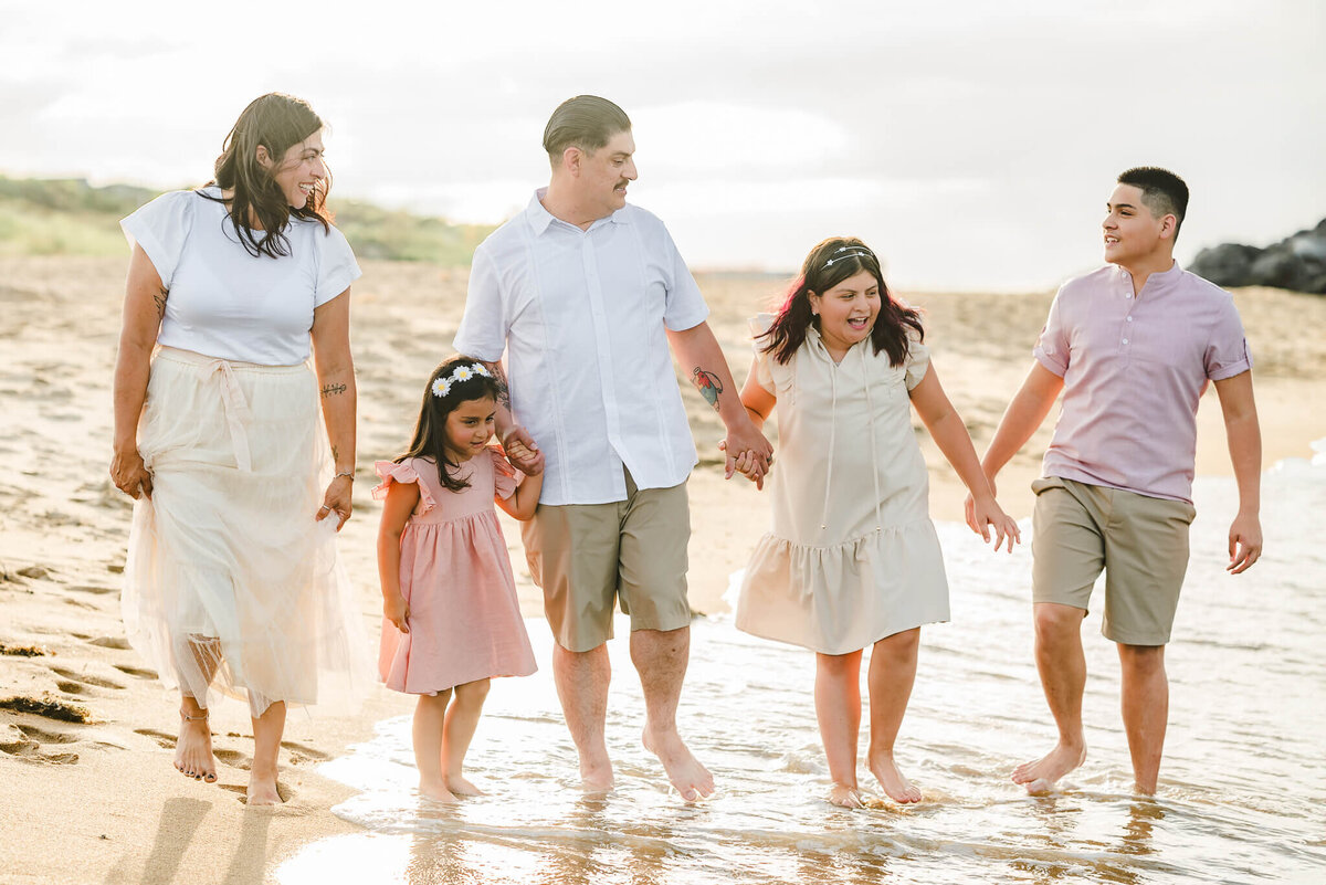 A family of five holds hands while they walk along s beach in the Outer Banks. The waves crash against their feet and the sun creates a glow behind them.