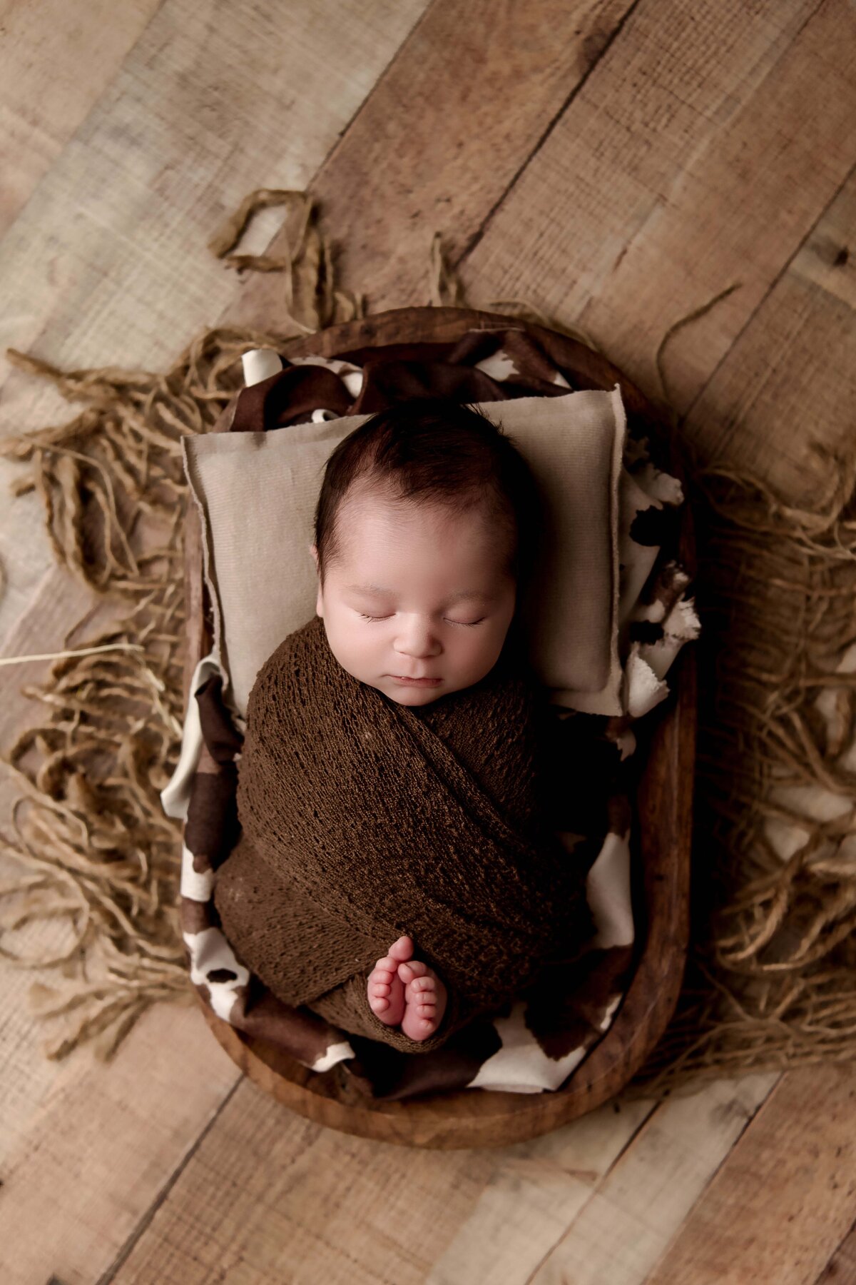 Newborn baby wrapped in a brown blanket, peacefully sleeping on a small wooden prop with textured fabric and wooden floor backdrop. His toes are peeping out at the bottom, posed carefully by professional newborn photographer in Mobile, AL