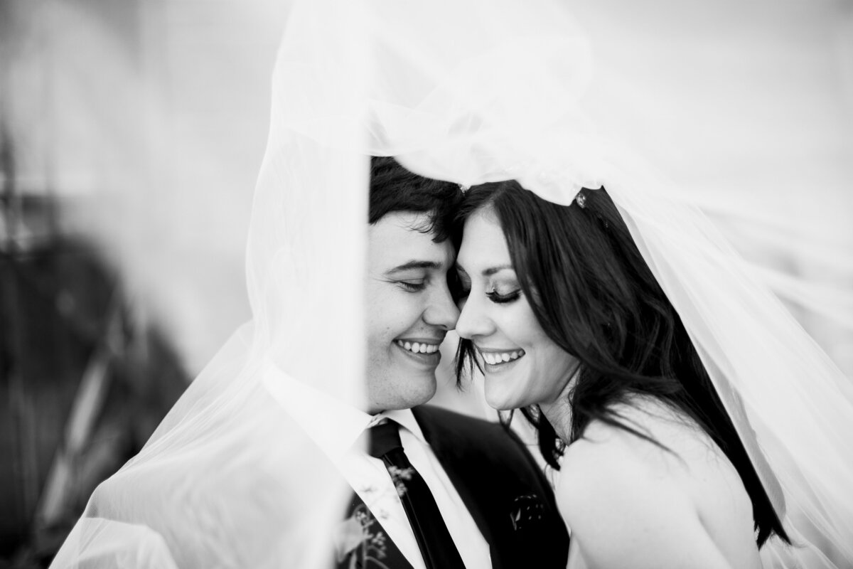 A black and white portrait of a bride and groom smiling and touching their faces together under the bride's veil which is framing their faces. The bride is on the right and is wearing a sleeveless, white dress, and long veil. The groom is on the left and is waearing a dark suit with a boutonniere.