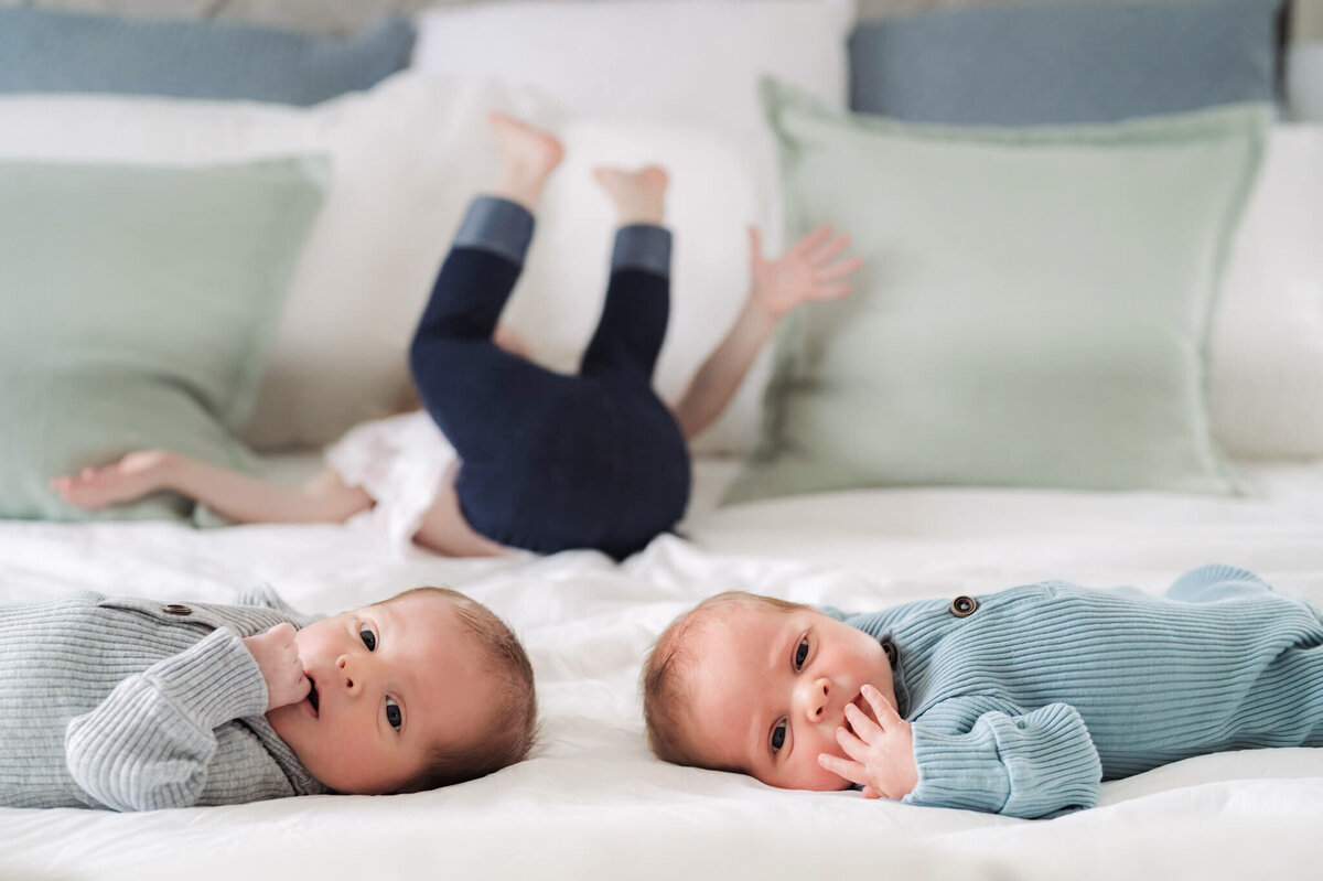newborn brothers look at the camera while their sister is blurred in the background with her legs up rolling over acting silly