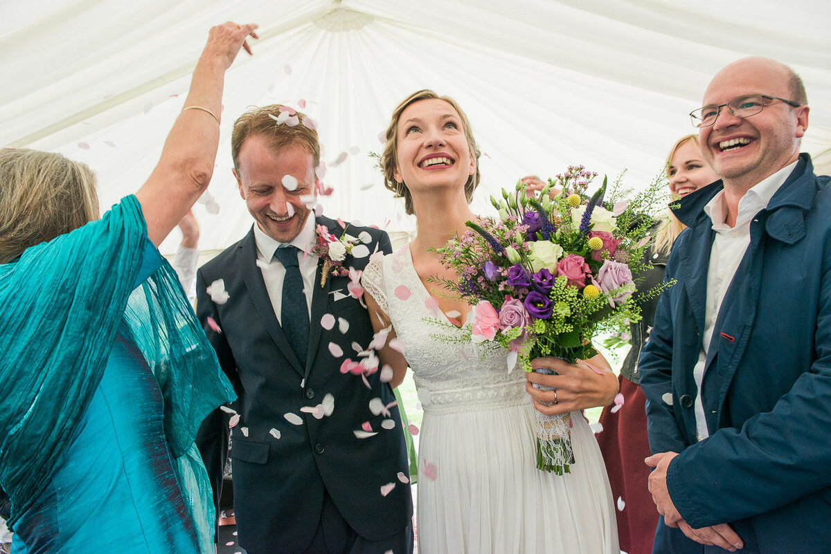bride wearing an empire style wedding dress, holding a colourful, wild flower bouquet walking down the aisle with her husband while confetti is thrown