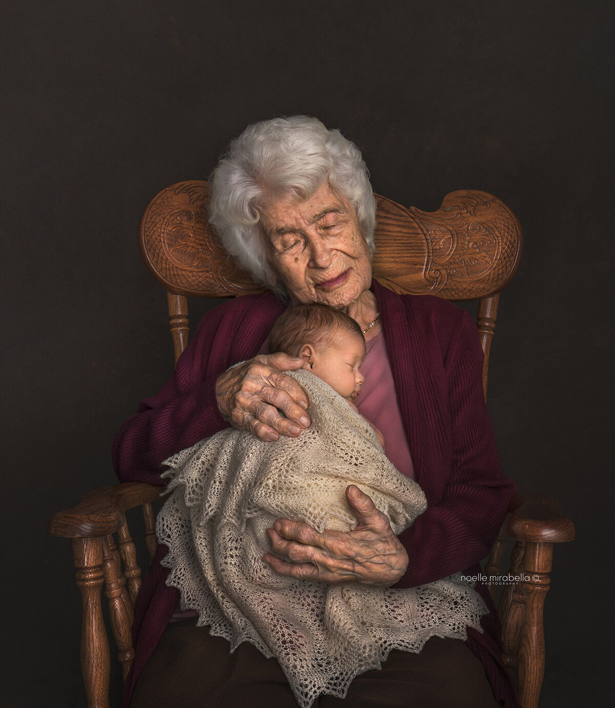 100 year old woman sitting in a rocking chair holding newborn baby. Pink and burgundy tones. Classic portrait.