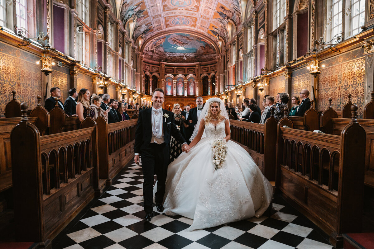 Bride and groom walk down the aisle at Royal Holywell Chapel in London, surrounded by guests in a richly decorated and historic chapel, showcasing an elegant and luxurious wedding celebration.