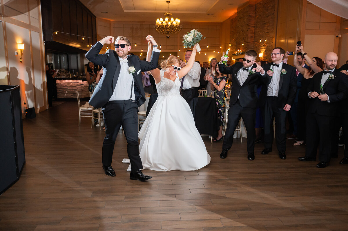 A bride and groom running into their wedding reception at the Madison in Riverside NJ.  Both are wearing formal wedding attire and sunglasses. They are holding their hands together in the air in celebration and they are welcomed into their wedding reception captured by South Jersey wedding photographer Allison McCafferty