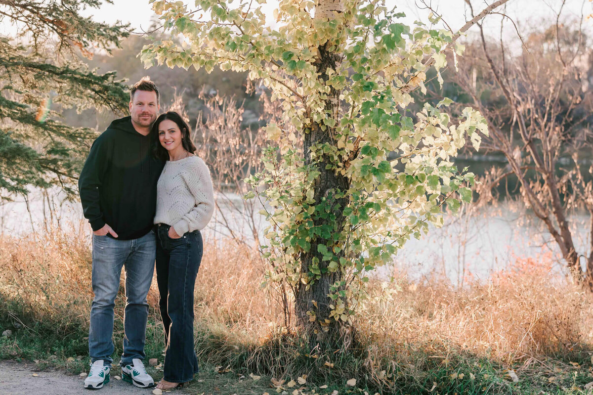 Mom and dad stand beside each other and near some trees and the river. 