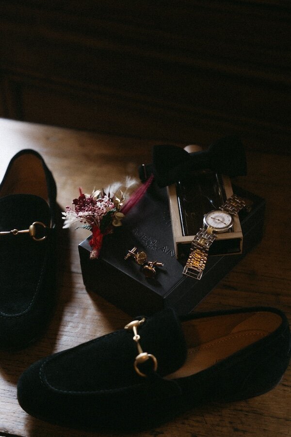 Mocassins en velours noir posés sur une table en bois, accompagnés d'une montre dorée, d'un nœud papillon et d'une boutonnière florale. Photo par Kamescop, photographe et vidéaste de mariage en Bretagne.