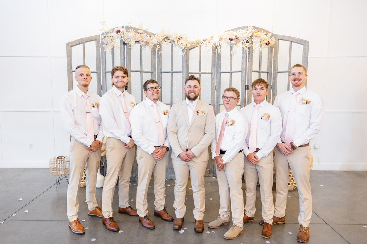 The groom stands with his groomsmen, all wearing matching beige and white attire with soft pink ties.