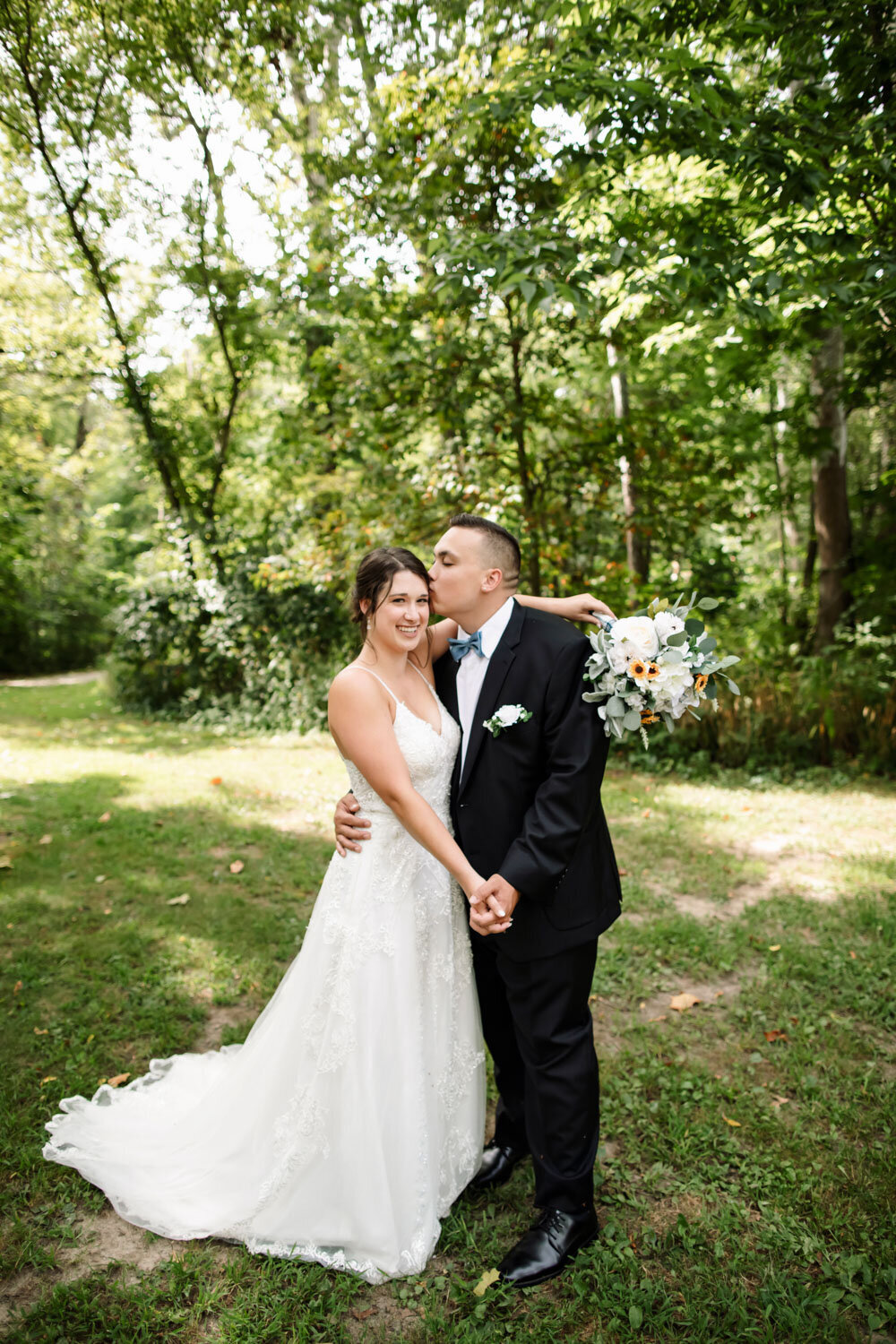 Groom kissing bride’s forehead while holding her close in a wooded setting