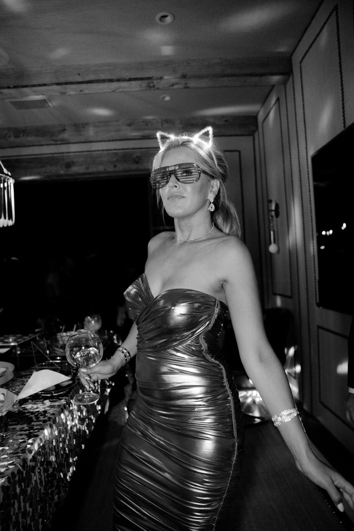 In a stunning setting in Virginia captured by photographer Elisabetta Marzetti and videographers Clayton Films, this scene unfolds with a person standing in a room, adorned in glowing cat ear headbands, futuristic sunglasses, and a strapless metallic dress. The individual exudes a festive aura, holding a beautifully decorated drink glass amidst a background of lively and celebratory decorations.