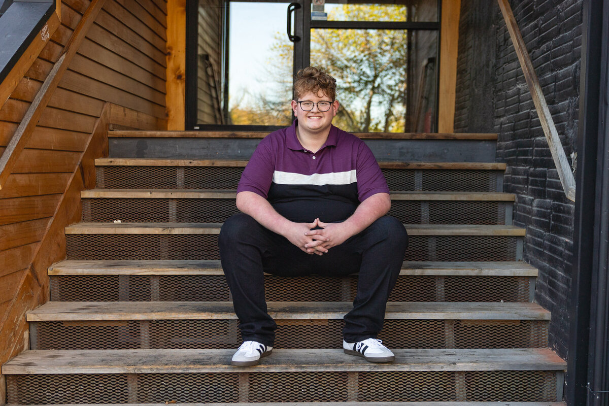 A senior guy is sitting on wooden stairs with hands together in Lawrence KS