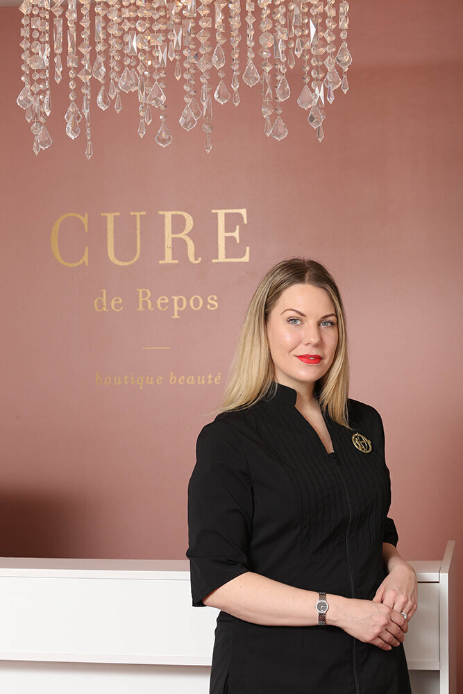 young blonde woman with red lipstick standing against a pink wall with CURE painted on the wall behind her