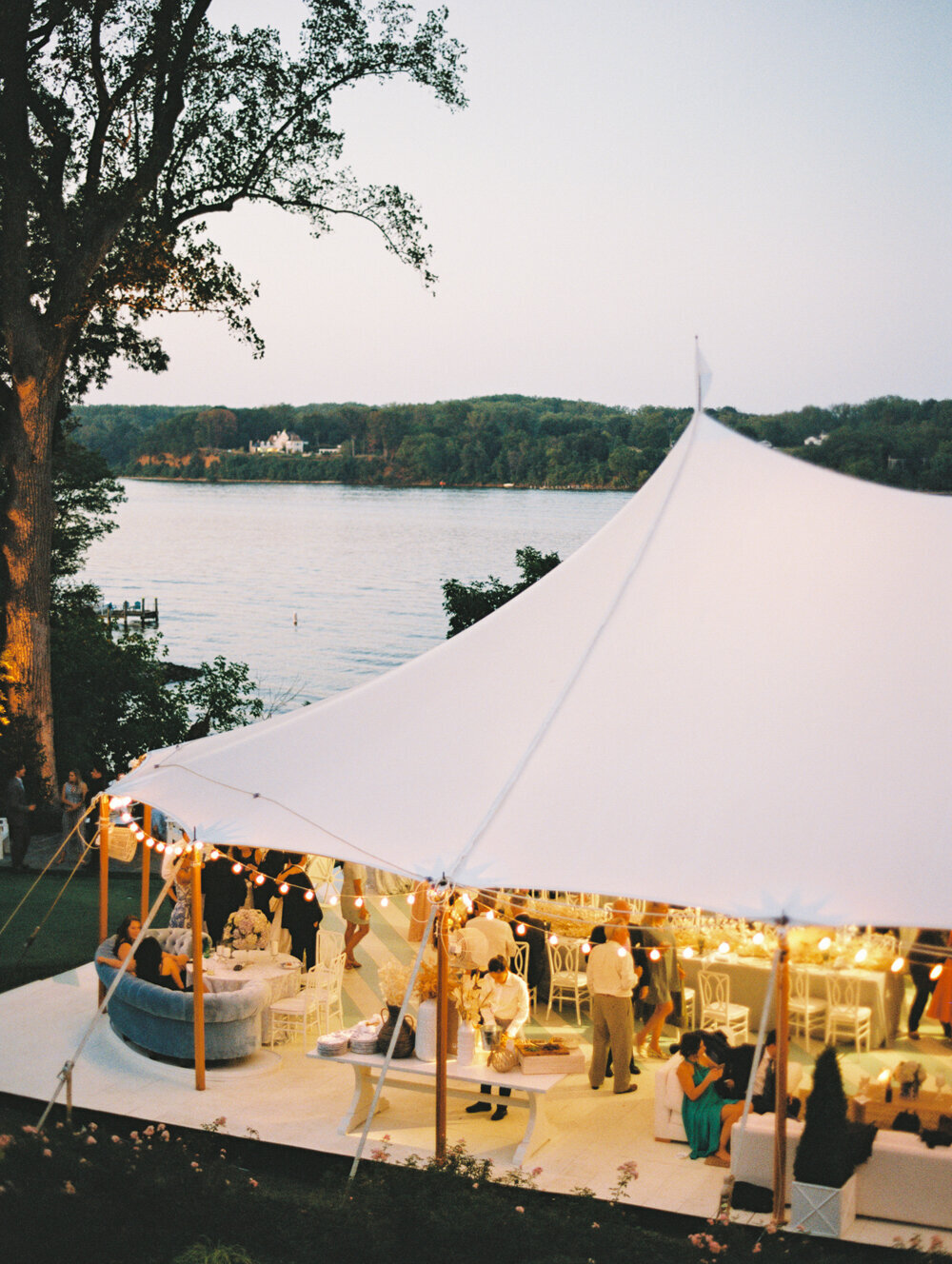 The photo was taken at The Hamptons, New York, a waterfront estate setting the perfect backdrop for a tented wedding captured beautifully by photographer Bonnie Sen. Videographers East West Films also expertly documented the event, ensuring all special moments were captured on film. This large white tent, set up elegantly for an outdoor event, overlooks a serene lake encompassed by lush wooded hills. Inside the tent, guests are seen mingling amidst the elegant setup featuring tables, chairs, and soft lighting that adds a touch of sophistication. The peaceful ambiance is further enhanced by the dusk setting, creating a truly magical atmosphere for the event.