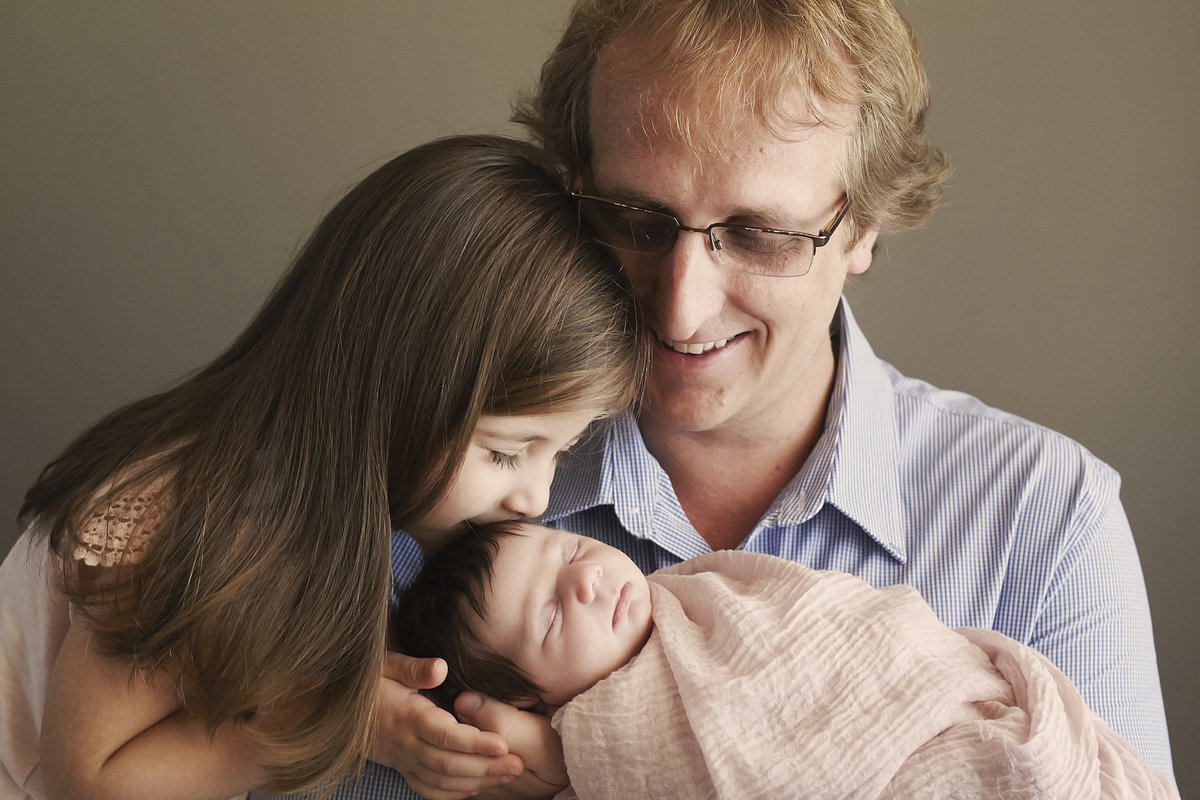 Dad holding newborn baby and little girl kissing the babies head and dad looking at the two