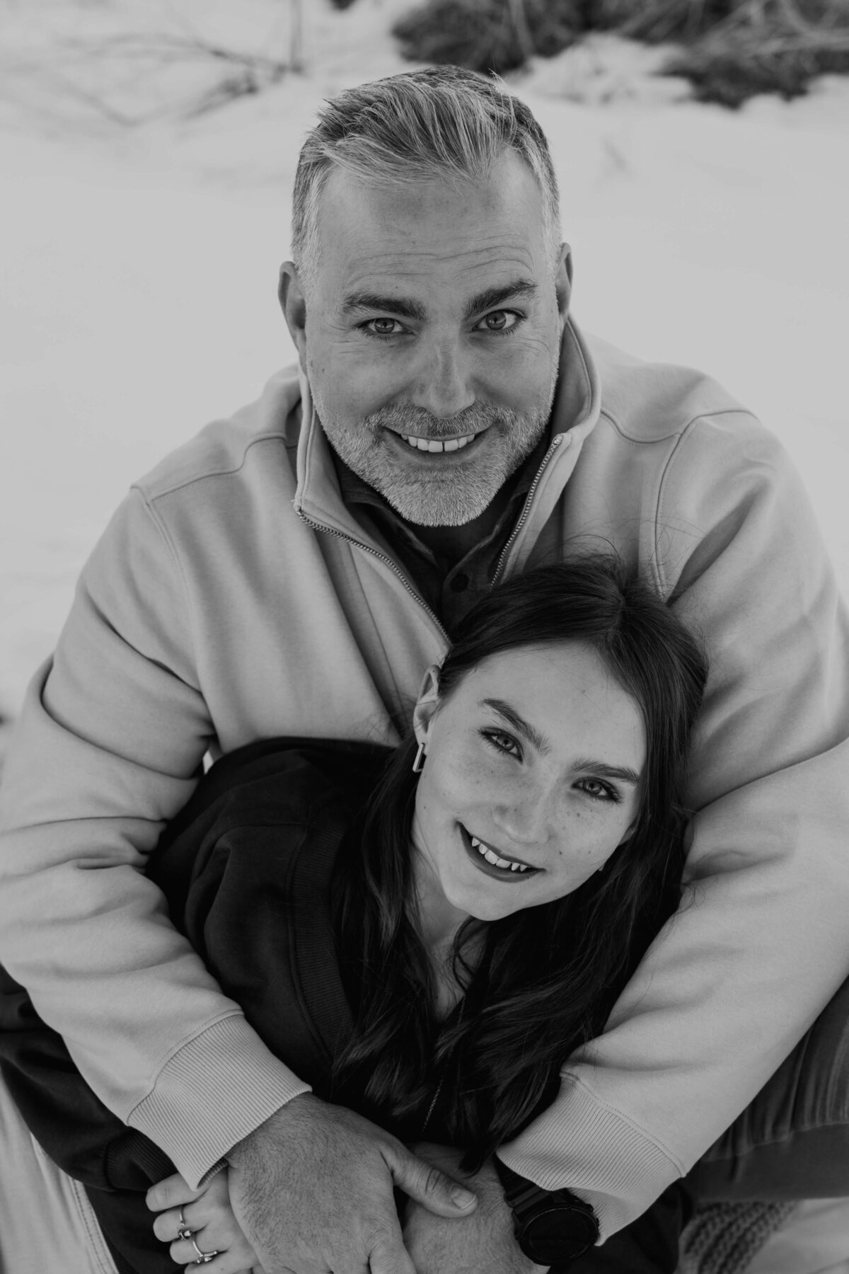 A beautiful, fine art portrait of a teenage daughter, laying in the lap of her dad, while they wear contrasting outfits with a snowy backdrop.