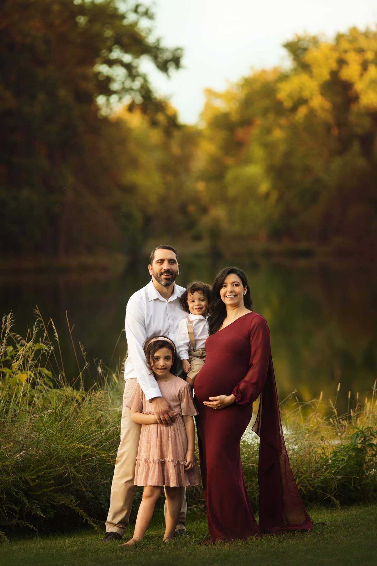 A happy family of four stand by a lake at sunset while pregnant mom holds her bump