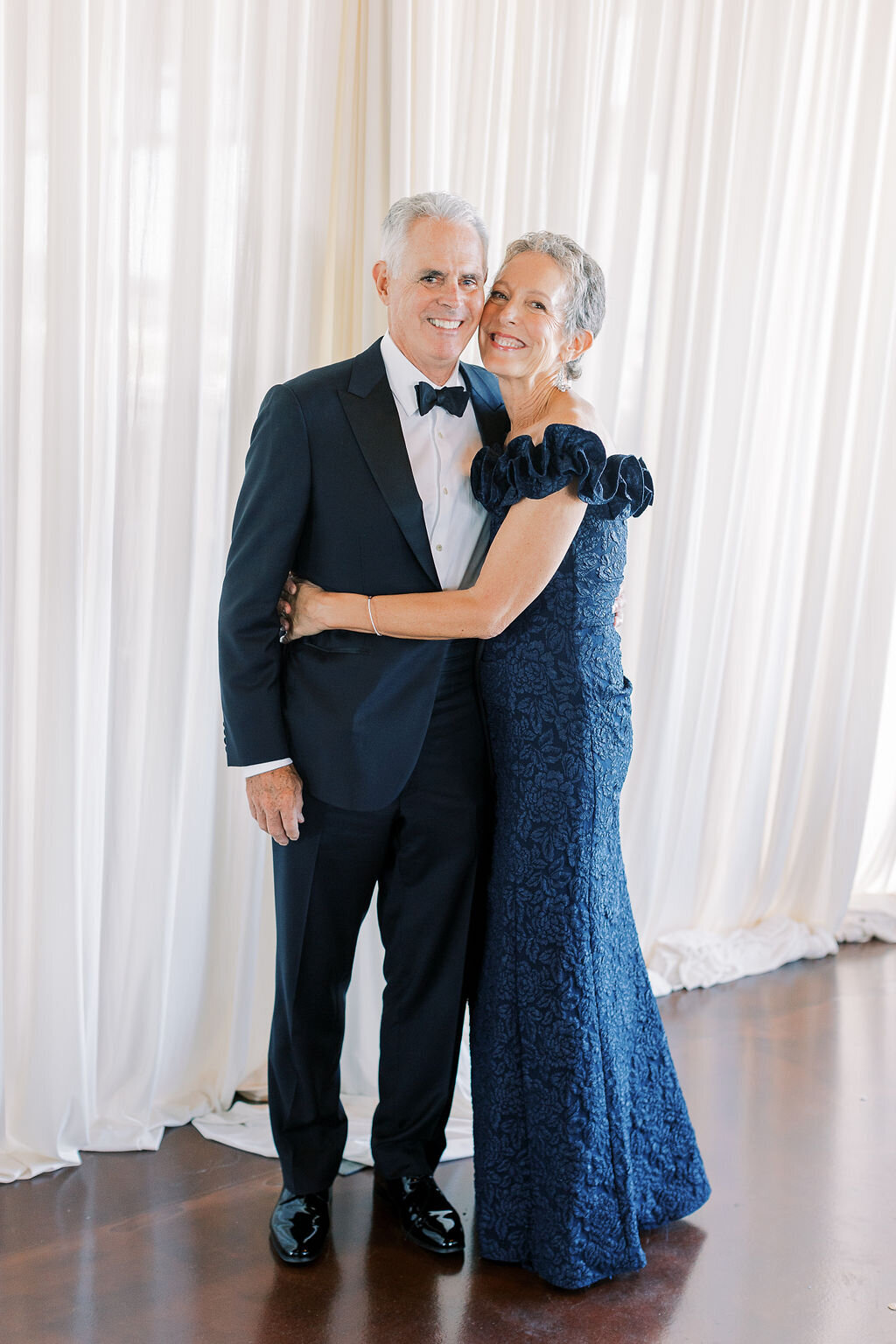 A couple dressed in formal attire poses in front of a soft draped backdrop.