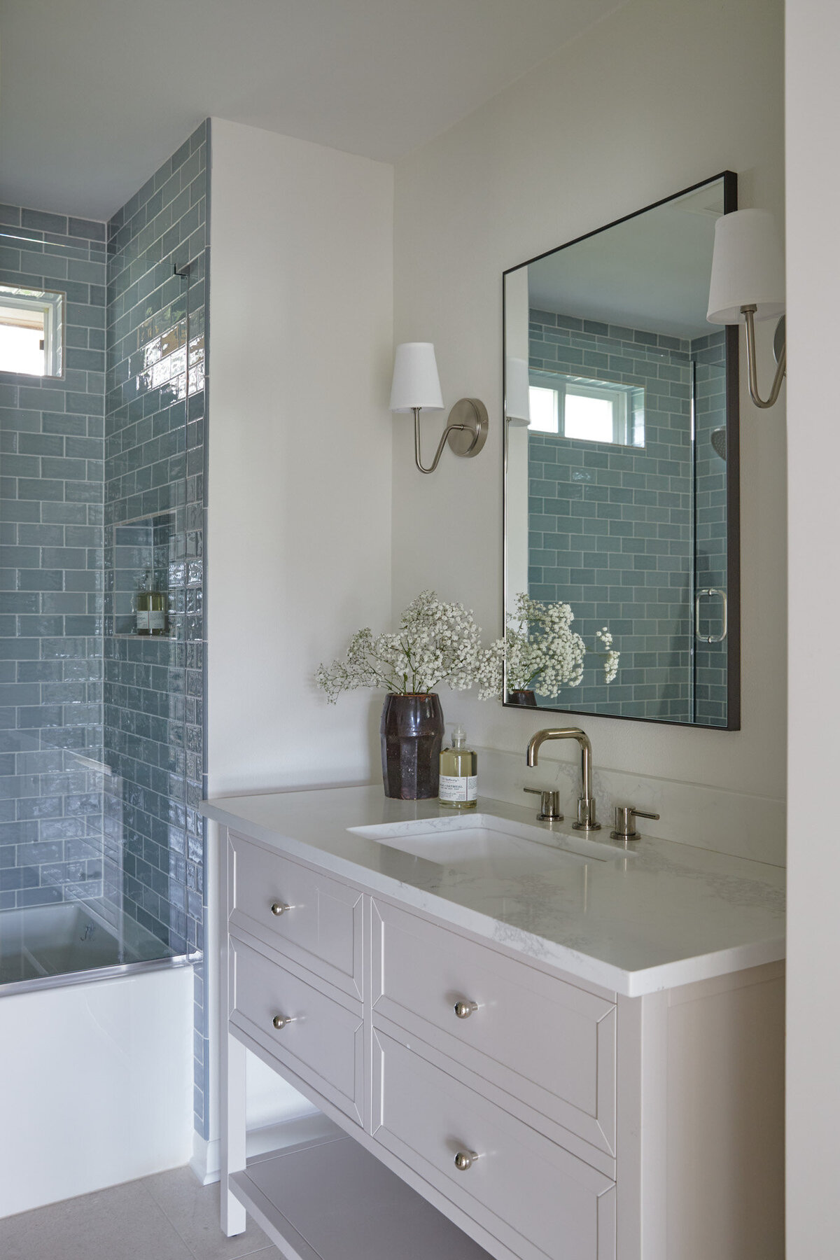 Bathroom with blue subway tile on the shower on the left. On the right is a single sink vanity in a neutral, beige paint with four drawers and an open shelf on the bottom. Above is mirror with a thin black frame and a silver sconces with a white shade on either side of the mirror.