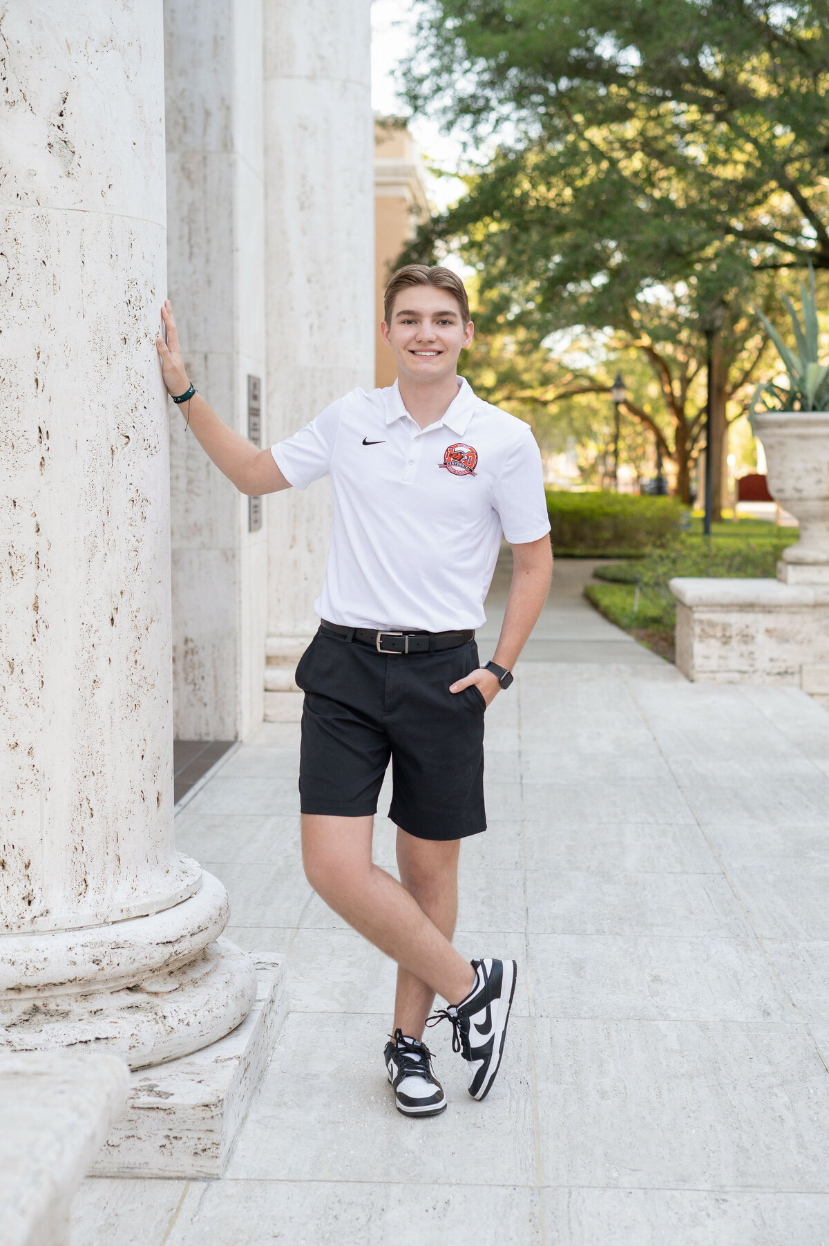High school senior boy holds pillar with one hand in pocket smiling.