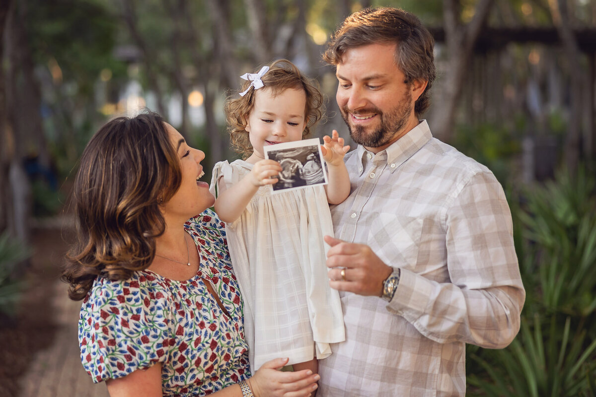 family of 3 with a little girl with curls holding a sonogram ultrasound photo about to be a family of four with a little boy.