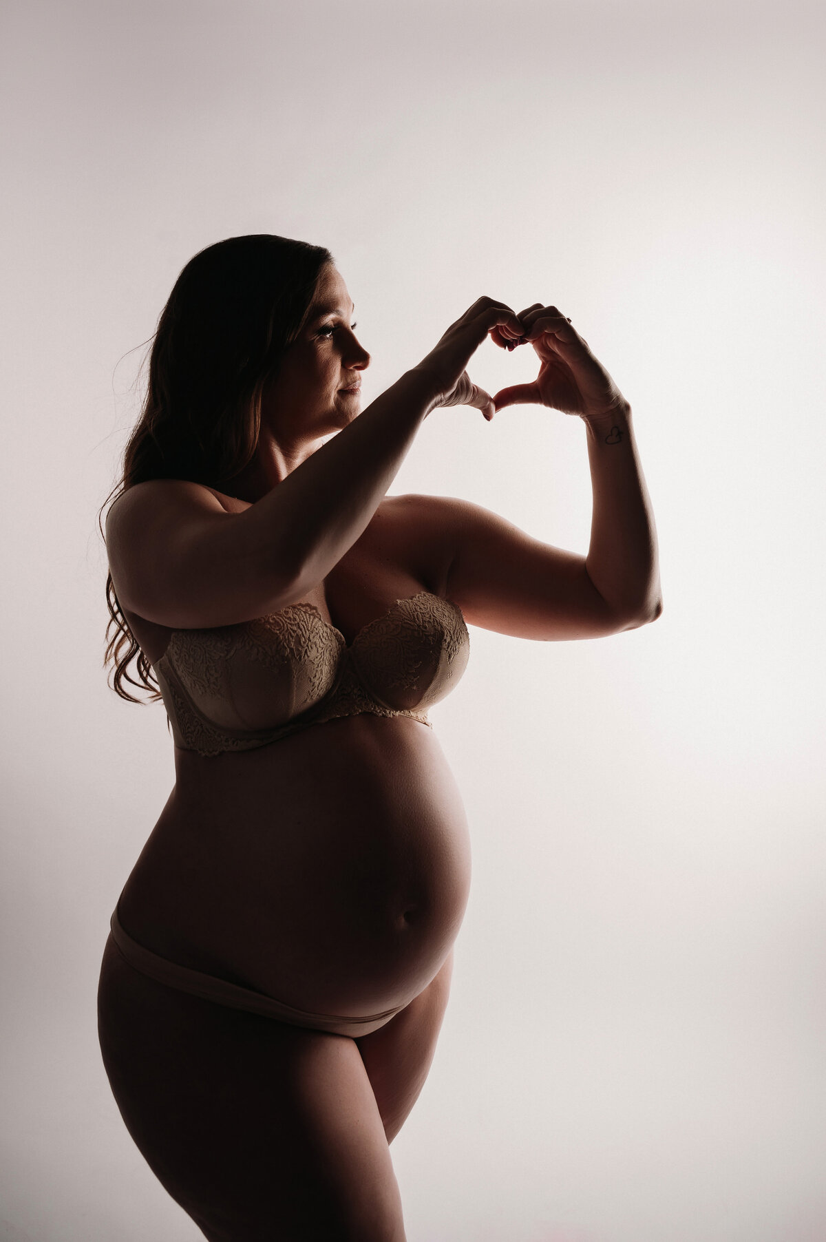 silhouette of pregnant woman's body making a heart shaper with hands in front of white backdrop
