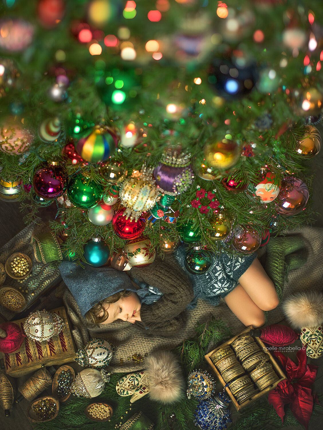 Small child sleeping under Christmas tree full of Christmas ornaments.