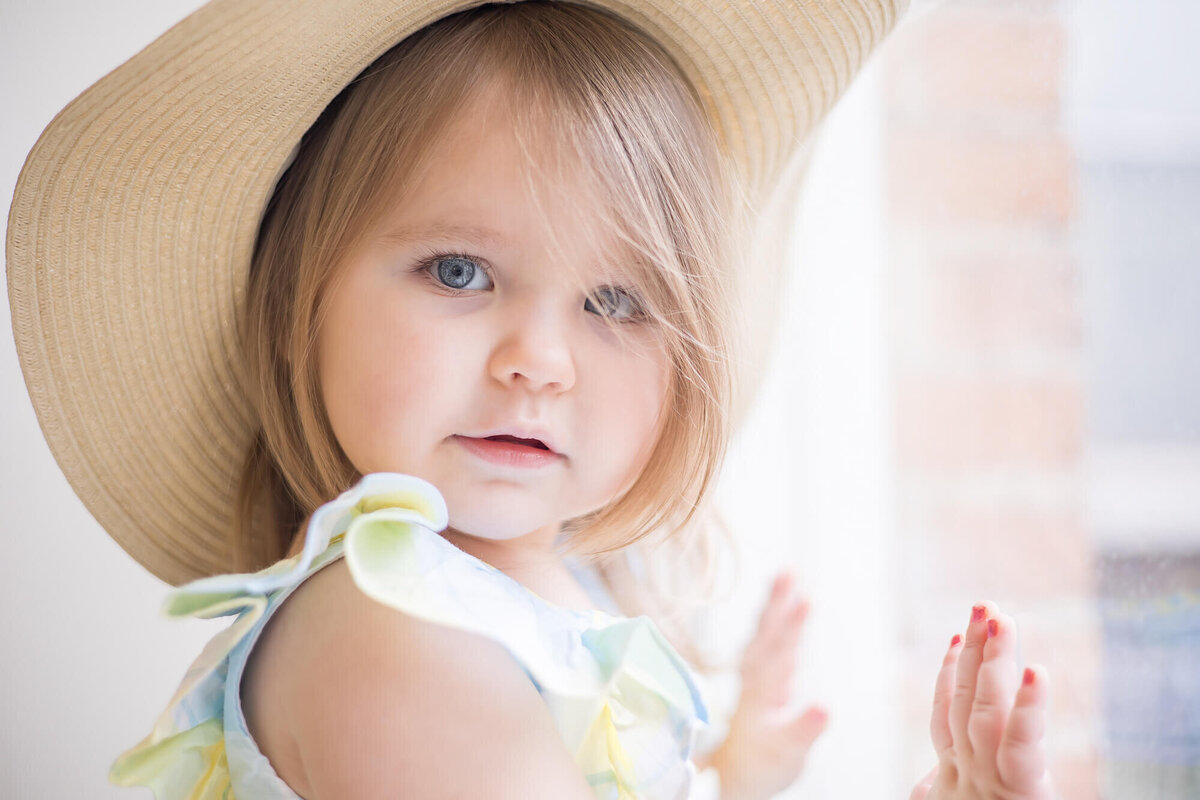 Stunning blonde two year old girl weraing a sun hat and putting her hands on a window