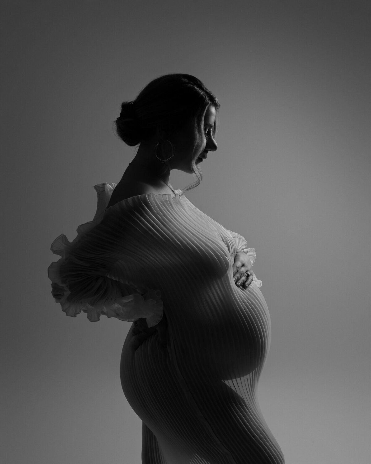 Black and white maternity photo in pleated fabric, captured with dramatic side lighting and a sculptural silhouette.