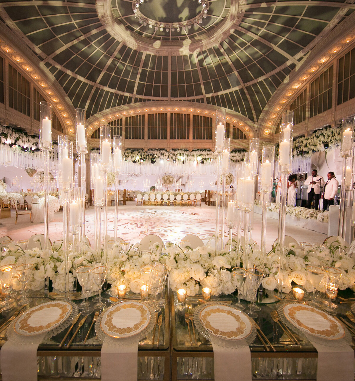 The stunning photo was taken at The New York Public Library in New York City by the talented photographer Christian Oth. It captures a luxurious wedding reception held in an opulent venue with a high, domed glass ceiling. The table is exquisitely adorned with white floral arrangements, tall candles, and elegant table settings. The sophisticated atmosphere is further elevated by the soft, ambient lighting, creating a truly unforgettable setting for the celebration. Videography for the event was expertly captured by East West Films.