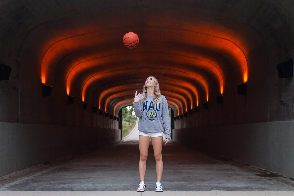 A person wearing a gray NAU hoodie and white shorts juggles a basketball under an illuminated tunnel with orange lights lining the ceiling. They stand centered, looking up at the ball.