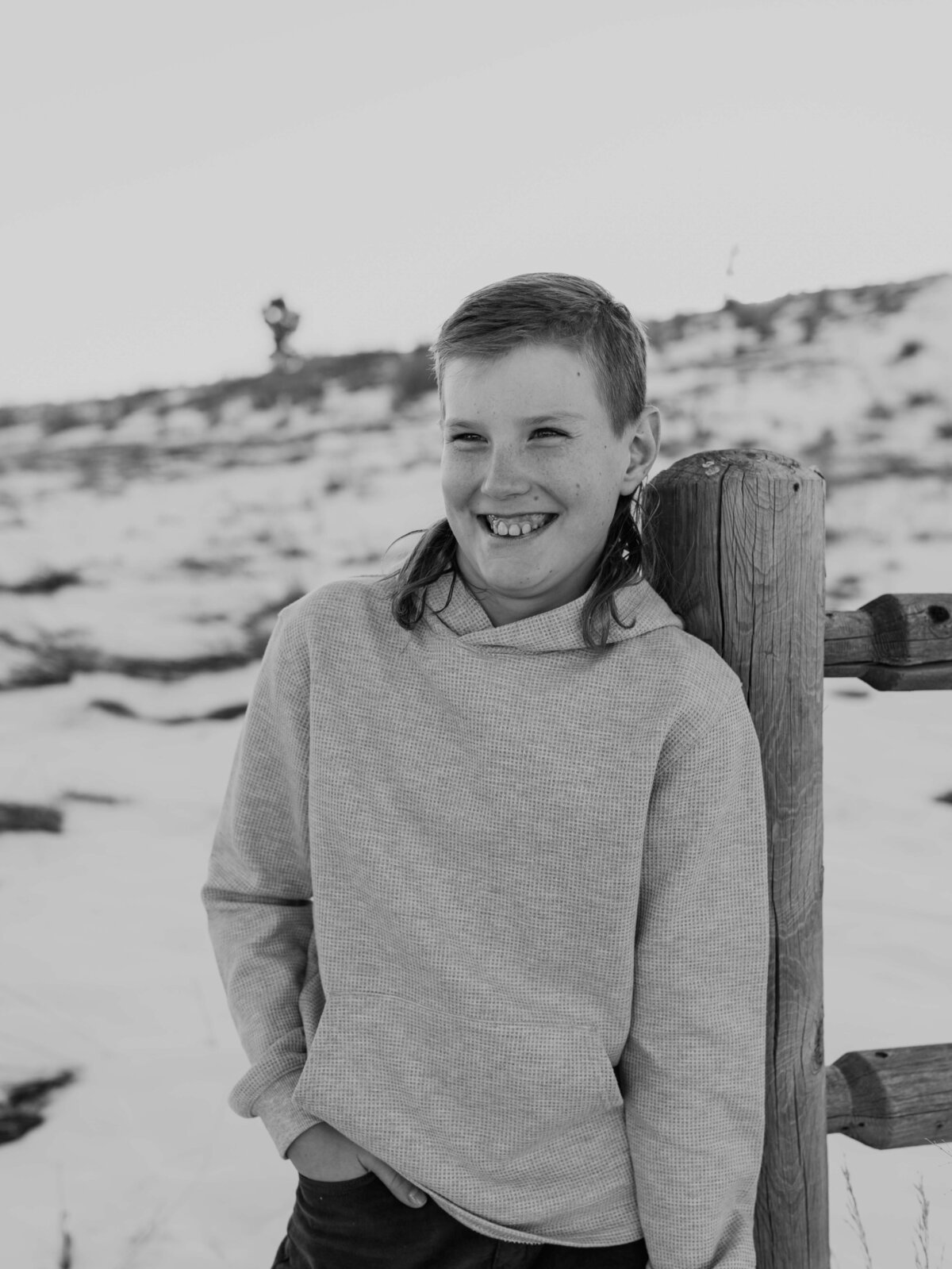 A young boy liens against a fence with ranch land behind him as he grins at the camera, wearing a long sleeve sweater and rocking a long hockey style mullet.