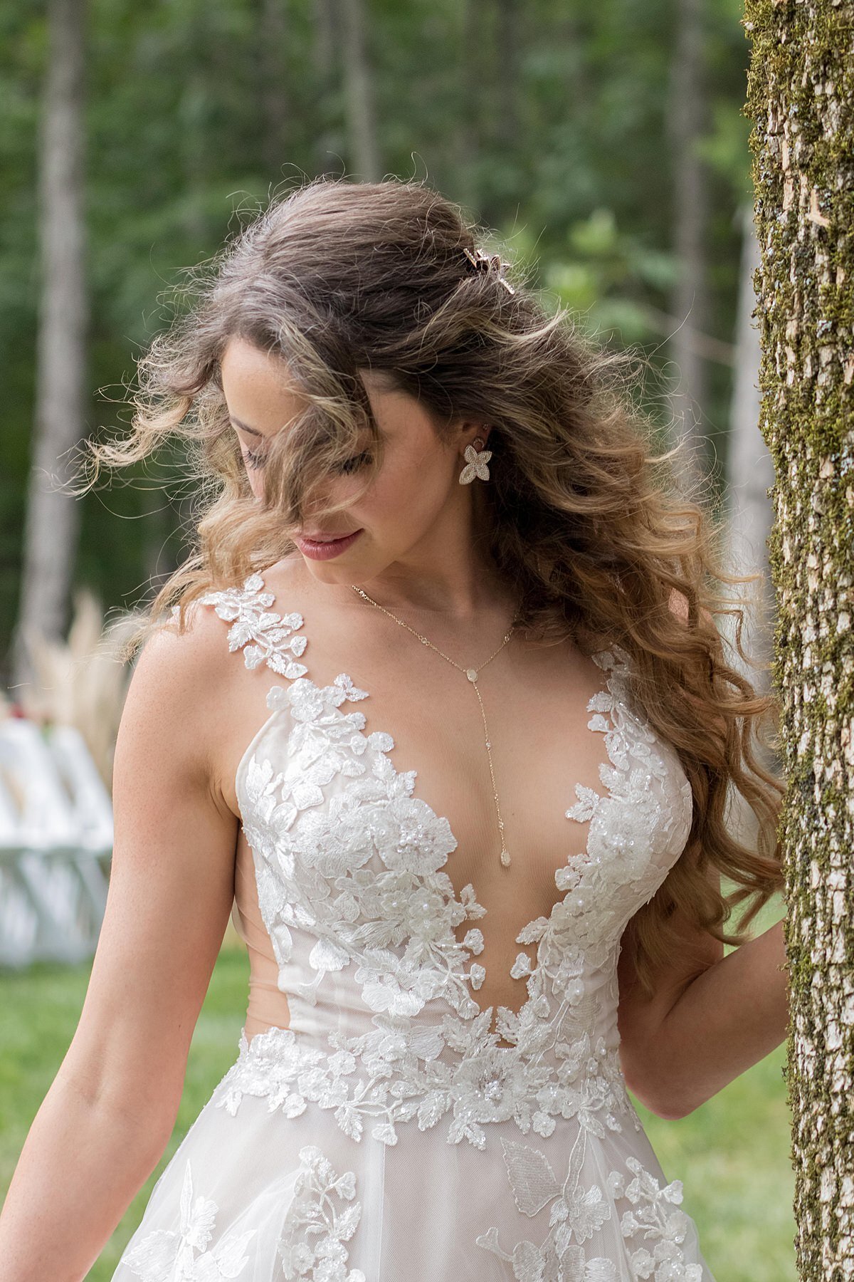 Bride wearing a delicate lace dress with a deep plunging neckline leaning up against a tree at her wedding ceremony at Saddle Woods Farm