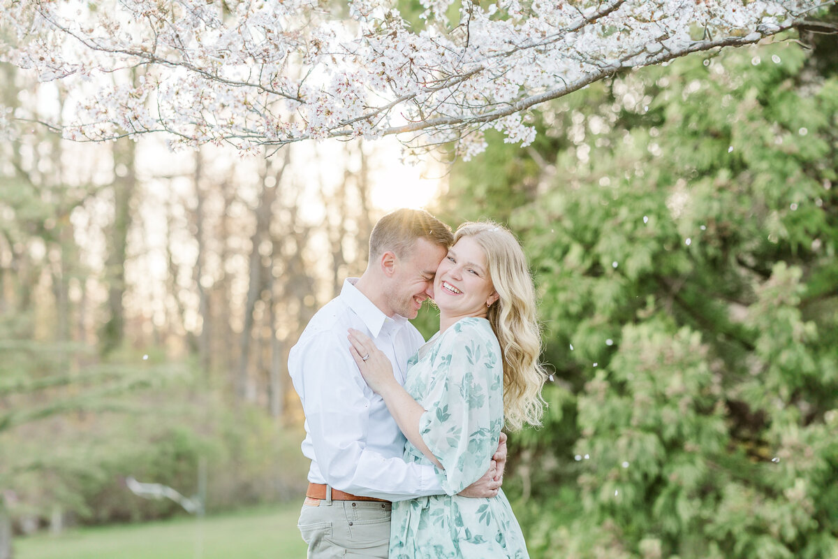 A newly engaged couple laughs while hugging under a blooming tree in a park at sunset