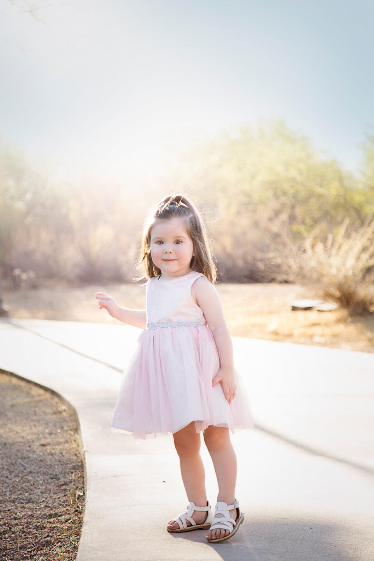 Toddler girl dressed in a light pink dress standing on a park path in front of dreamy sunset during her session with las vegas milestone photographer Jessica Bowles