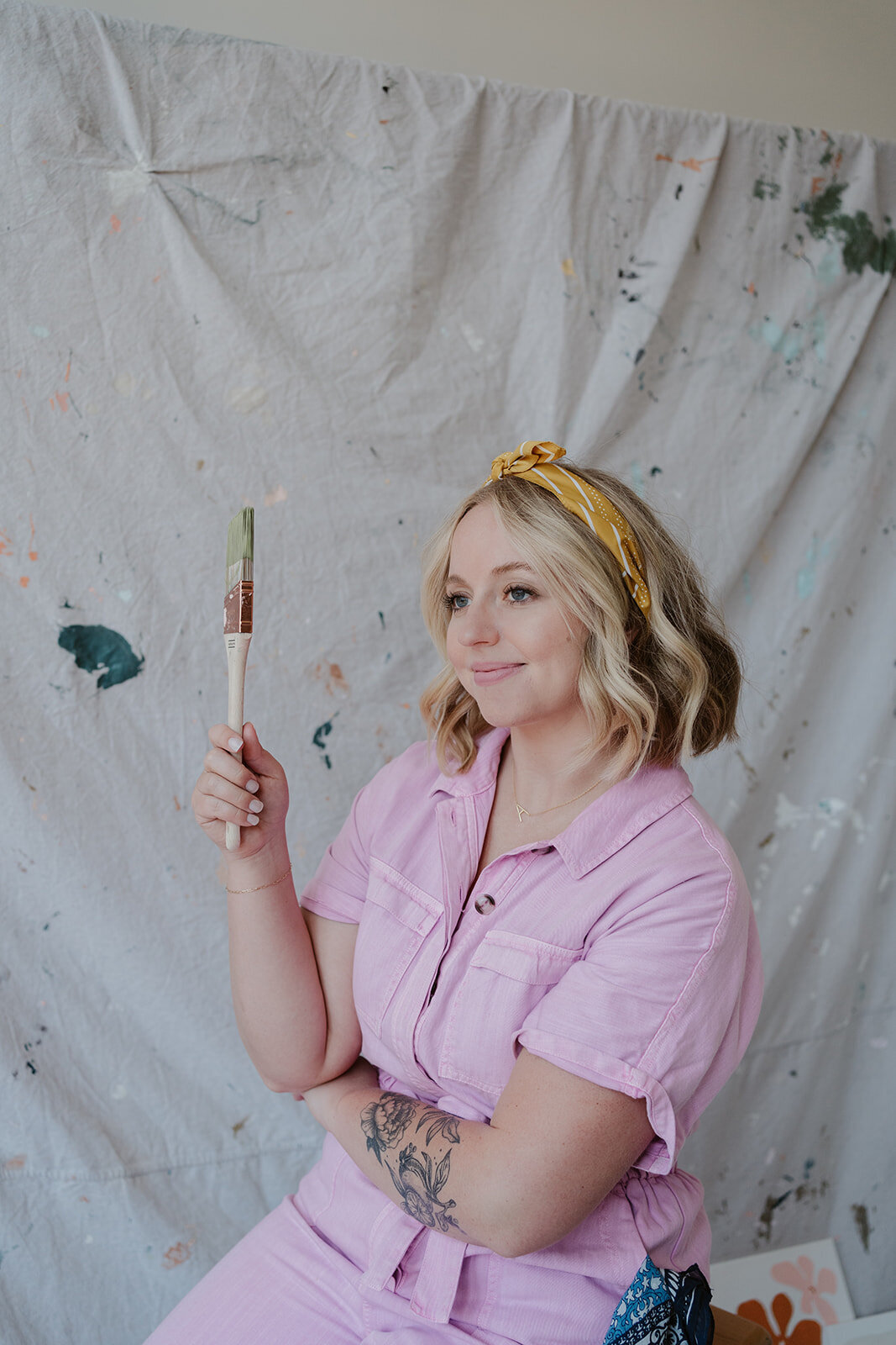 Woman in a pink jumpsuit holding a paintbrush and posing in front of a painted backdrop during a Kalamazoo studio branding session.