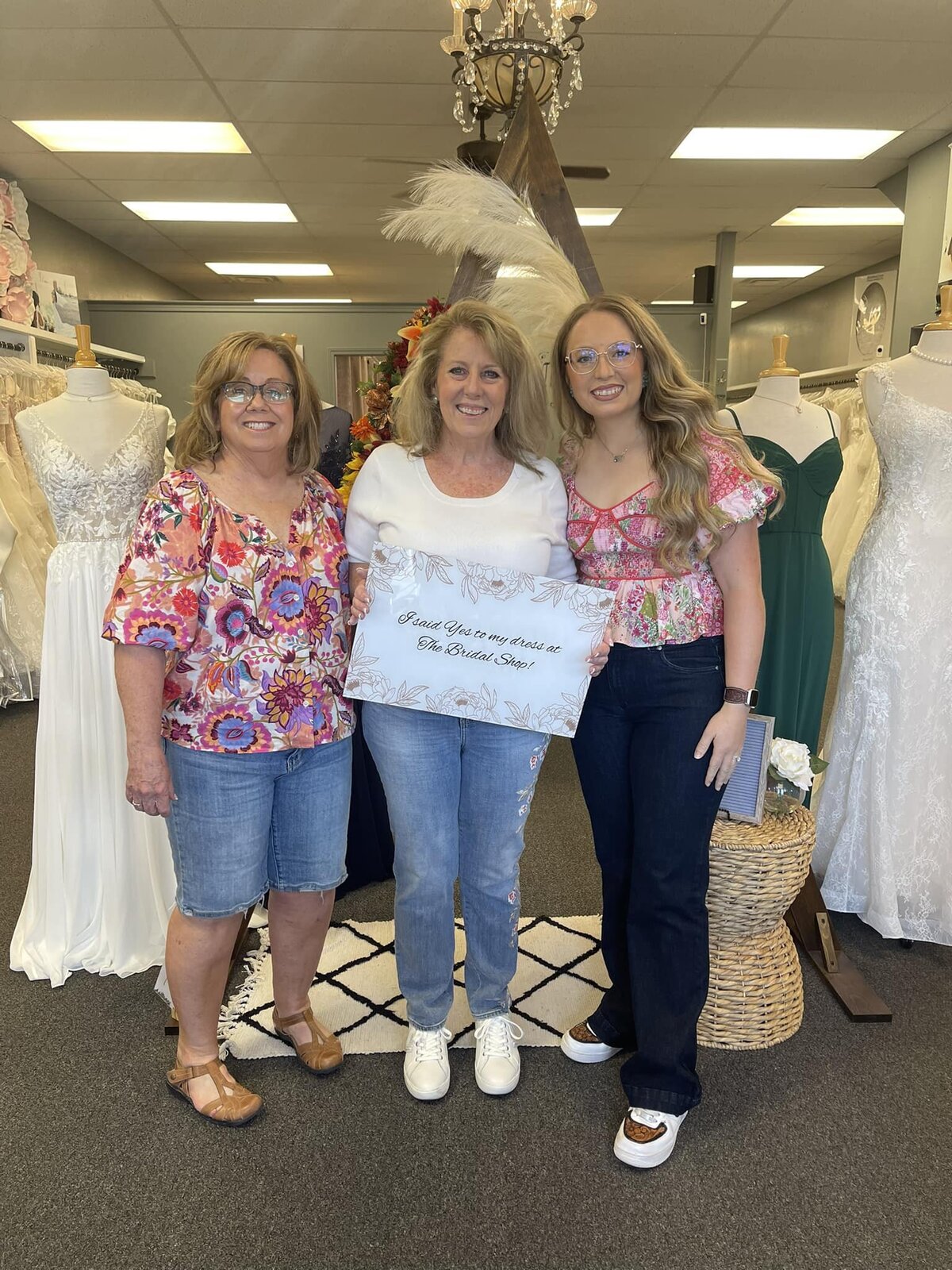 Three women standing in a bridal shop holding a sign announcing a dress purchase