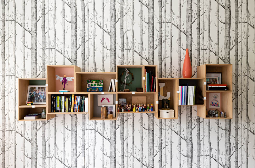 Custom shelving with kids' books and decor is set against Cole & Son wood wallpaper in a modern new construction home in Hingham, blending style and function.