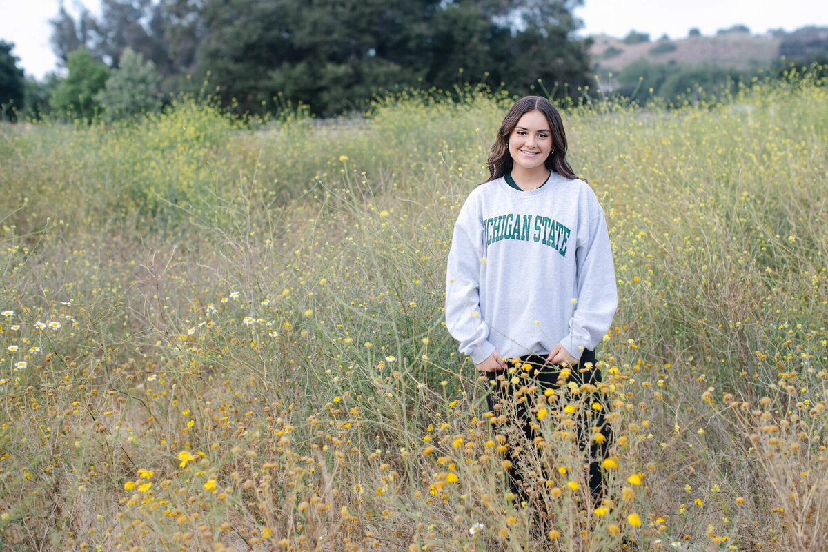 A person wearing a Michigan State sweatshirt stands in a field of wildflowers. The background features tall green grass and distant trees under an overcast sky.