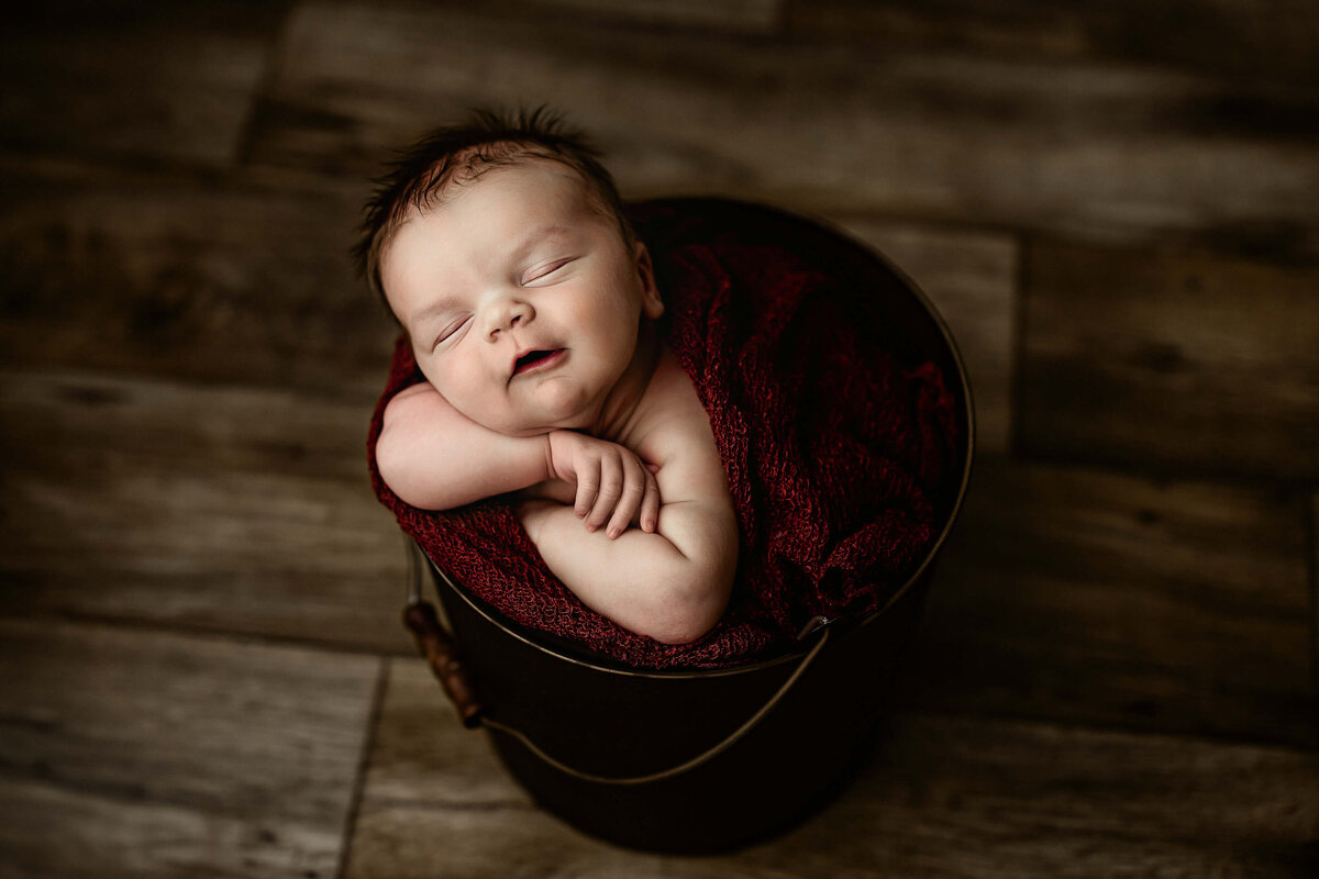 A sleeping baby is snuggled in a dark bucket, wrapped in a maroon blanket, resting peacefully on a wooden floor background.