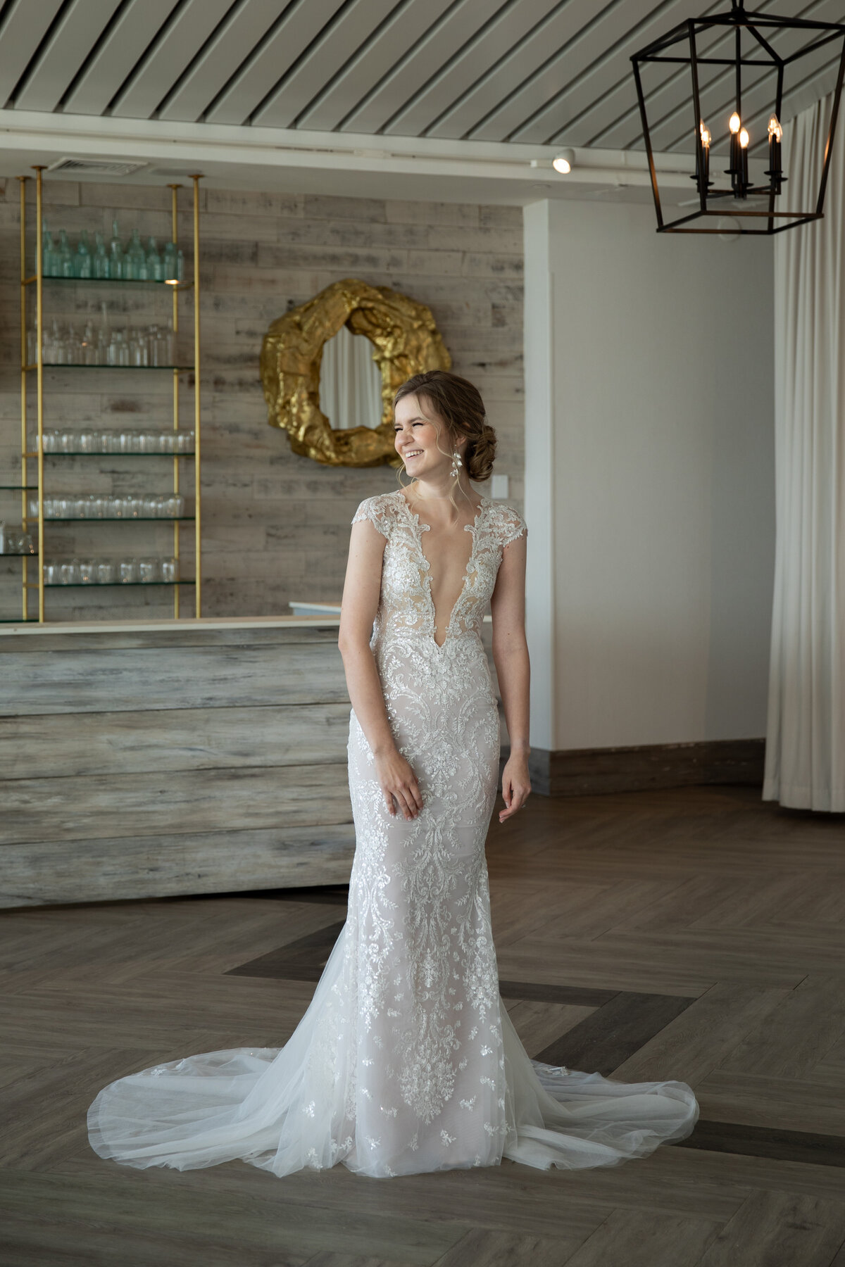 Wedding Photographers in RI | Kelsey Sheehan Photography Timeless Rhode Island Weddings | A smiling bride in an elegant lace gown stands indoors, with rustic wooden decor and glass shelves in the background, conveying joy and elegance.