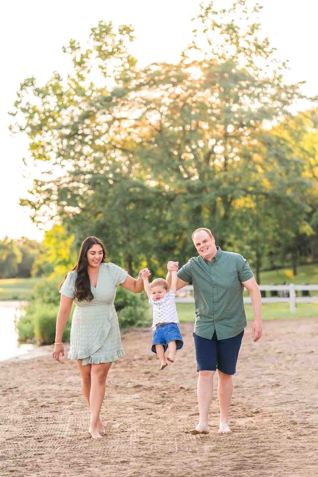 mom and dad walking on the beach at Kensington Metropark Michigan holding toddler boys hand and swinging him back and fourth as he laughs