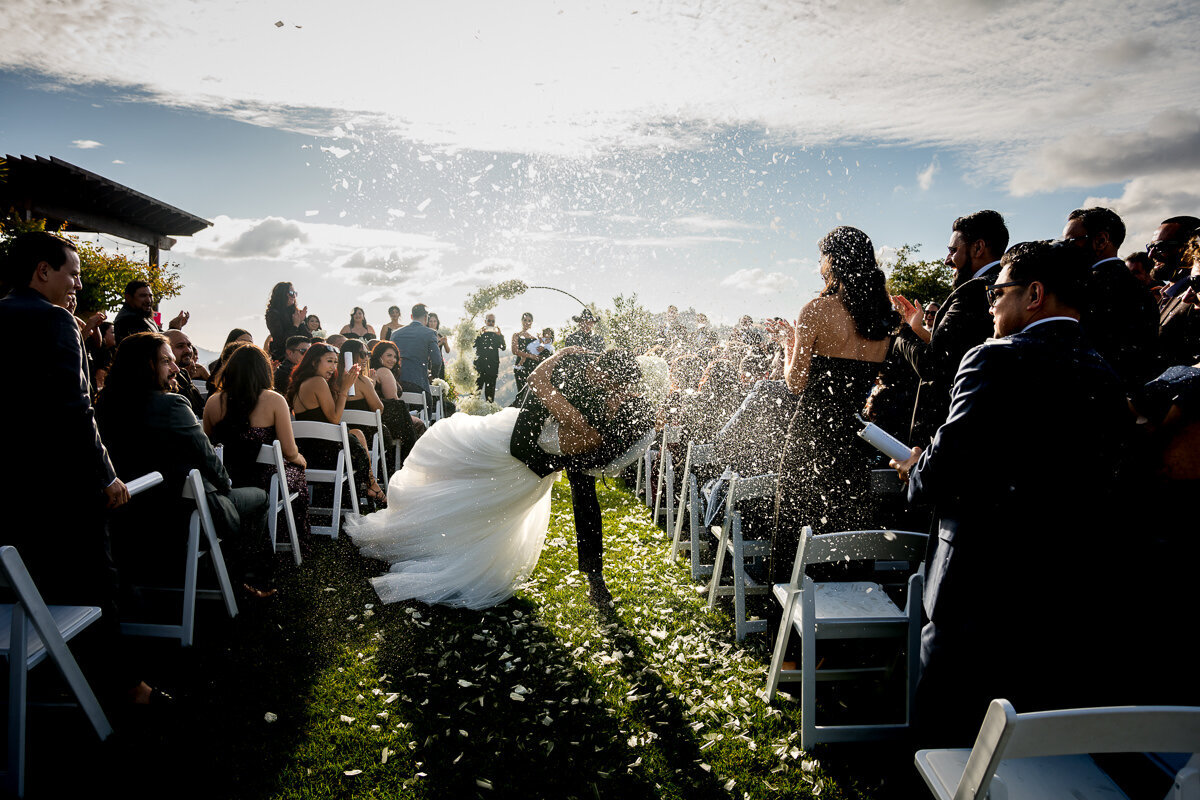 A picture-perfect moment as the newlyweds share a romantic kiss while walking down the aisle, surrounded by a shower of vibrant confetti, capturing the joy and celebration of their special day.