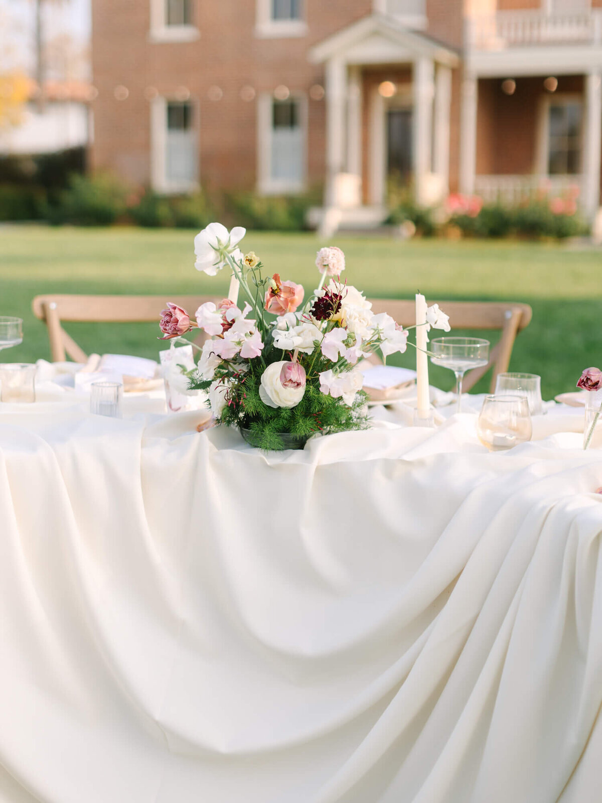Elegant outdoor table setting with white tablecloth, floral centerpiece of pink and white flowers, candles, and glassware. 