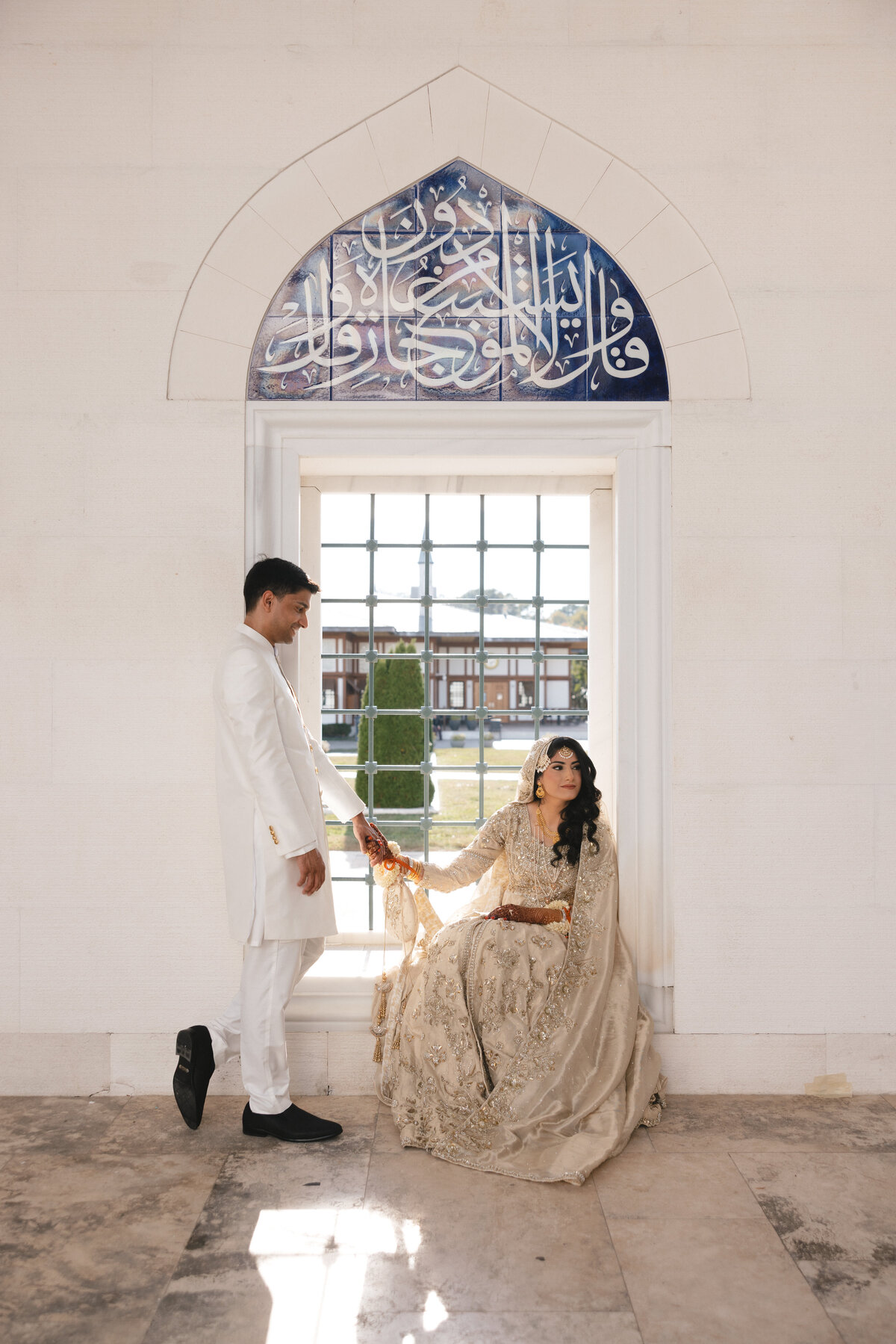 Bride and groom in gold and white traditional attire posed in a bright architectural alcove with Arabic script overhead.