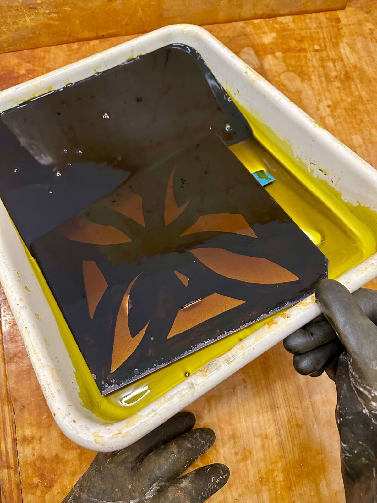 A gloved hand holds a copper plate partially submerged in a yellow acid bath, with floral shapes masked off in black resist.
