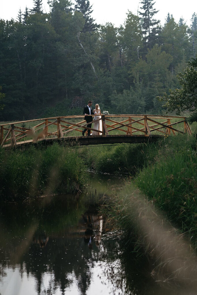 Elegant bride and groom at wooden bridge at The Valley Weddings, a rustic and majestic wedding venue in Westerose, Alberta, featured on the Brontë Bride Vendor Guide.