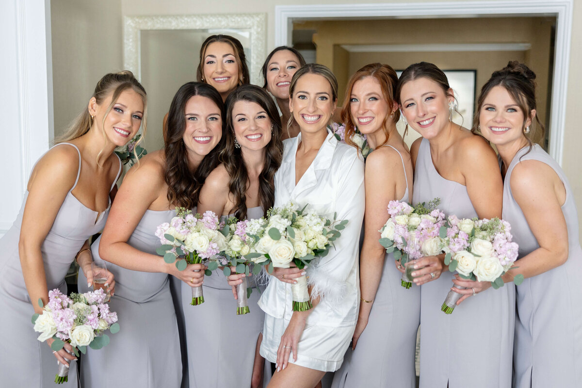 A bride in a white dress is surrounded by eight bridesmaids in light lavender dresses, each holding a bouquet of white and pink flowers. They are smiling and standing closely together in a room with a light-colored interior.