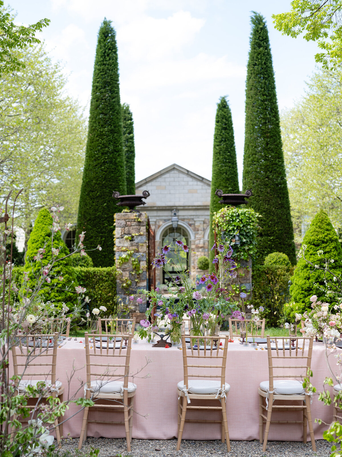 An outdoor garden wedding reception setup featuring a long table with a blush pink tablecloth, gold Chiavari chairs, and lush floral arrangements, framed by manicured greenery and a classical stone structure in the background.