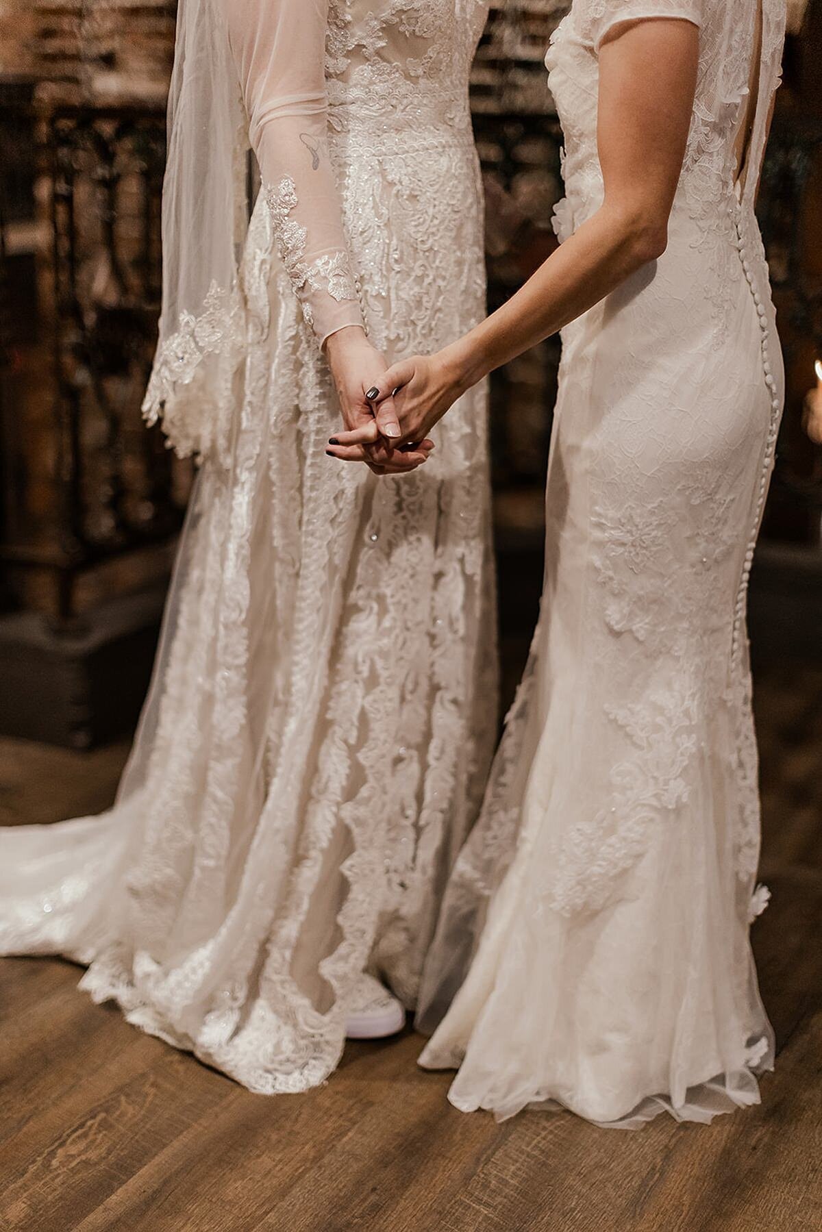A bride wearing an ivory long sleeved lace wedding gown and veil holds hands with a bride wearing a white wedding gown with a lace overlay and tiny buttons down the back.
